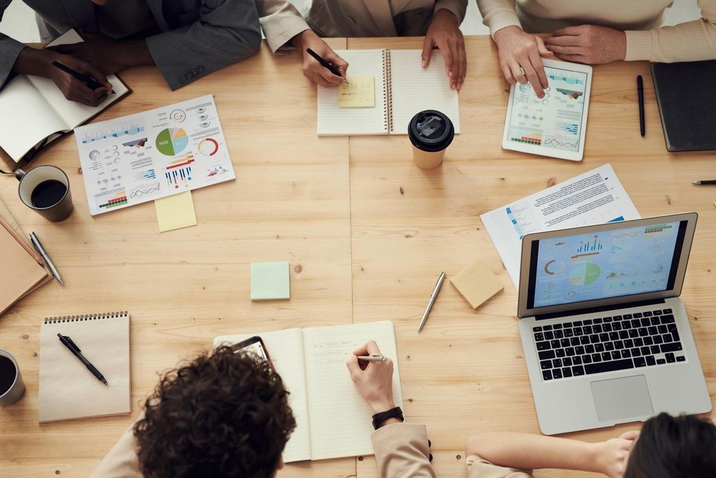 Ein Team von Geschäftsleuten sitzt an einem großen Holztisch in einem Meeting. Auf dem Tisch liegen Notebooks, Tablets, Notizblöcke, Diagramme und Kaffeebecher. Mehrere Personen machen sich Notizen oder analysieren gemeinsam Daten und Statistiken auf digitalen Geräten. Die Szene vermittelt eine produktive Arbeitsatmosphäre mit Fokus auf Business-Analyse oder Projektplanung.