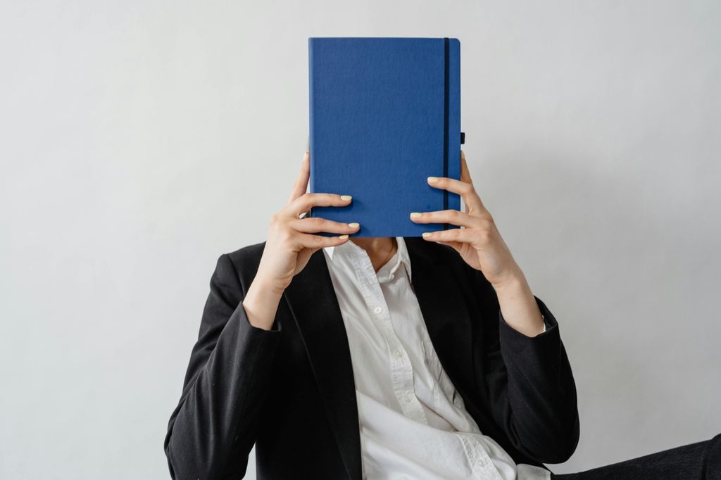 Eine Person in Businesskleidung (schwarzer Blazer, weißes Hemd) sitzt vor einer hellen Wand und hält ein geschlossenes, blaues Notizbuch vor ihr Gesicht, sodass dieses vollständig verdeckt ist. Die Fingernägel sind dezent gelb lackiert.